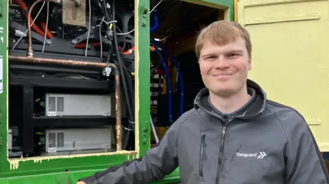 A man in a grey top and with brown hair is looking at the camera. He is next to equipment on the left of the photo.