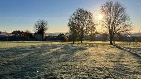 WeatherWatchers/BrizzleDrizzle A field at dawn with a low sun creating long shadows of the few trees in view, over the frosty grass