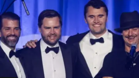 Getty Images Donald Trump Jr (left to right), JD Vance, Charlie Kirk, and Kid Rock on stage all smiling, wearing tuxedos except for Kid Rock who is wearing black and a black hat.