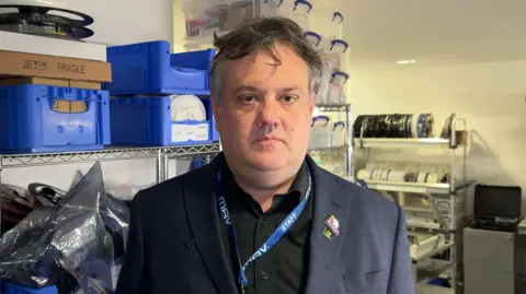 Chris Shepherd looking at the camera with a neutral expression. He is pictured in a store room with lots of items on shelves. He has grey hair and is wearing a black shirt and blue suit. He has a bright blue lanyard around his neck.