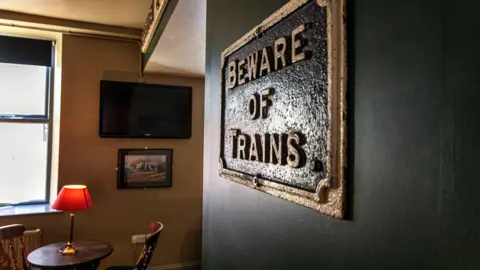 Keighley and Worth Valley Railway A sign on the wall of a pub reads "beware of trains". A table and chairs with a red lamp is in the background.