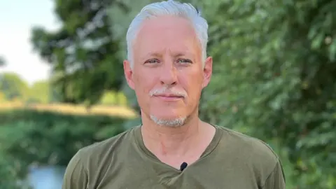 Pete stands in the middle of the picture with a light green "Angling Trust" t-shirt on. Behind him the River Pang and some trees are visible.