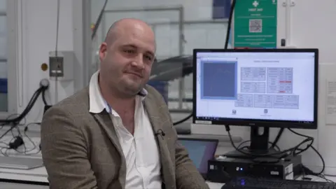A bald man looks side on of the camera, he is wearing a brown jacket with a white shirt. Behind him is a lab and a data set on a screen.