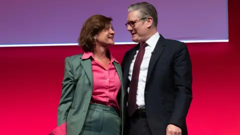 Getty Images Eluned Morgan stood with Sir Keir Starmer at Labour conference, with a red background behind them. Morgan is in a green jacket and trousers and a red shirt.