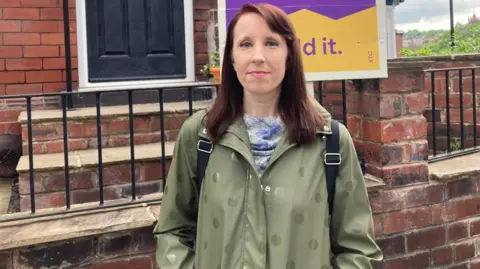 Simon Thake A woman in a green overcoat stands in front of a house with a sold sign