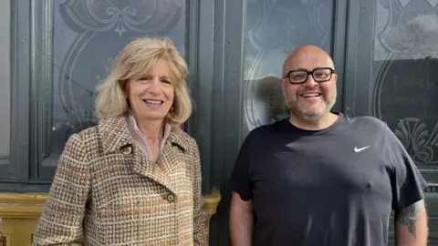 Lili Sheppard/BBC Gemma Dempsey and Jim Barber smiling for the camera. They are stood outdoors beside some pub windows.