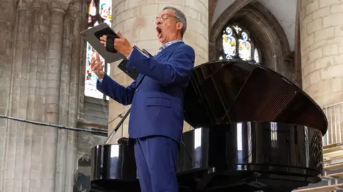 James O'Driscoll Roderick Williams performing a song recital inside Gloucester Cathedral. He is wearing a blue suit and holding a music book. He is stood in front of a piano with stain glass windows in the background. His mouth is wide open as he sings passionately, he is holding a device in his hand which he is reading from.