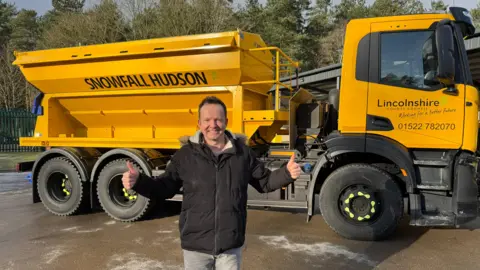 Paul Hudson standing in front of a big yellow gritter. He is wearing a black coat and grey jeans and is looking at the camera, holding his thumbs up. The gritter has "Snowfall Hudson" written in black block letters on the side