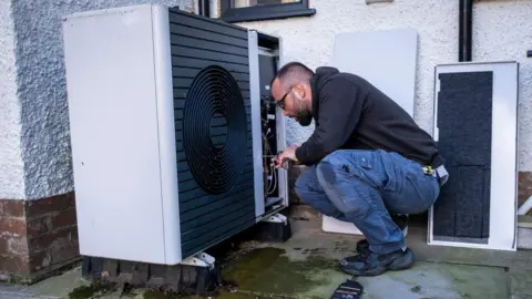 Getty Images A man crouches down to carry out work on a heat pump, which is fixed to the ground outside a property. He is wearing a black hooded top, denim combat-style trousers and black trainers. He has cropped dark hair, a beard and is wearing glasses.