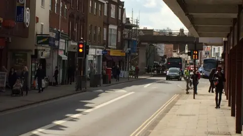 A view down Magdalen Street in Norwich, showing cars, buses, traffic lights, pedestrians, and shop fronts. A road bridge goes across the street in the distance.
