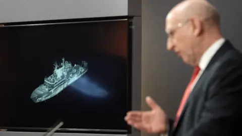 Getty Images Britain's Defence Secretary John Healey, shown with a screen displaying an image of Russian ship Yantar in the background
