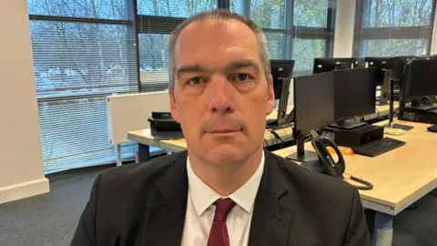 Norfolk Police Phill Gray is seated in front of several computer screens, with a window visible in the background. He has short gray hair, is clean-shaven, and wears a black suit paired with a crisp white shirt and a bold red tie. His gaze is directed straight at the camera, giving a composed and professional impression.