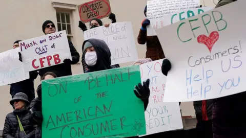 Reuters A group of protesters holding signs with slogans including "Musk and Doge out of CFPB" and "On behalf of the American consumer" 