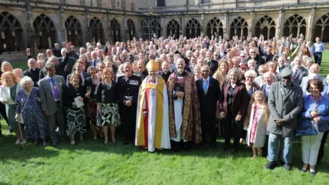 NNP View of more than 100 men and women on the grassed central area with the open walls of the gallery behind them. At centre and front of the crowd are the bishop and a dean in their clerical wear. Other people are formally dressed, mainly in sober colours. Some are holding their hands in the air and most are smiling. It is a sunny day.