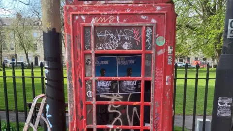 A red phone box covered in graffiti. It is in front of a green space.
