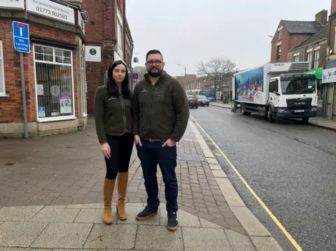 A man and woman standing on a pavement next to a high street