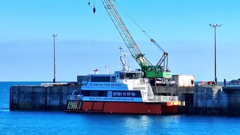 The Dart Fisher ferry docked in Alderney, painted red and white with the Alderney Ferry Services name and phone number on the side and a crane operating behind