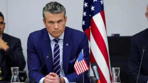 Reuters a seated Hegseth in blue pinstripe jacket with blue tie striped in pink and American flag handkerchief, leans forward toward a black microphone looking stern with a small American flag before him to his left on the table and a large US flag behind him, also to his left