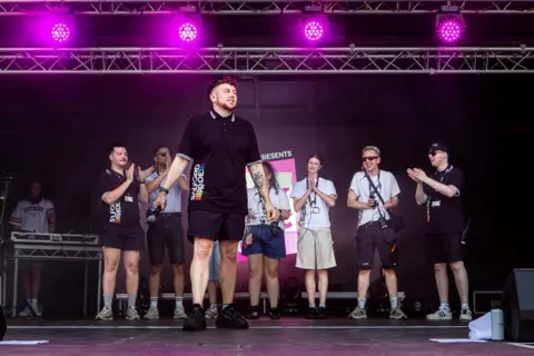 OhMyMedia / Olly Rigby Reece Holmes, event lead, at the front of a stage holds a microphone while a group stands behind applauding under purple lights