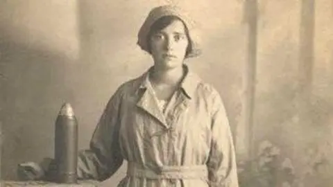 Gloucestershire Family History Black and white photograph of a woman, thought to be that of Gloucester Canary Girl Nell Slatter. She is wearing the working overalls and hat of the National Filling Factory No. 5 which was established at Quedgeley. There is a large shell cartridge sitting on a table next to her.