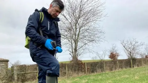 A man is standing in a field wearing navy water proof trousers and a coat. He is holding a device used for the sampling process.