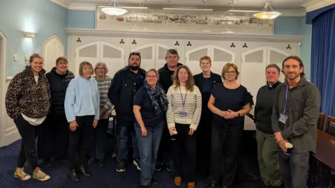 A group picture of a dozen or so individuals standing in a room with a blue carpet and blue and white walls.