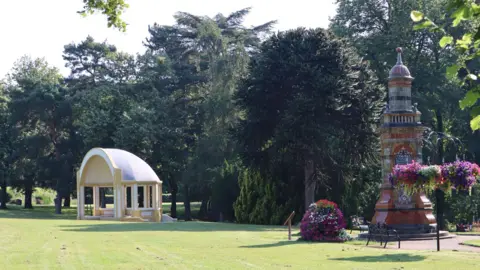 Brinton Park in the sunshine with a monument and a bandstand area. There are hanging baskets and flowers in a pot and trees in the background.
