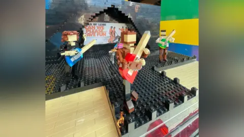 Brad Charlton-Wells A small black lego sculpture depicting a stage. On the stage there are four lego people - on the left, a guitarist with brown hair and blue guitar, on the right a guitarist with a green guitar and black hair. At the back is a drummer with a pink shirt on. At the front of the stage is a man with light brown hair and a red guitar. On the back of the stage is a sign that reads "kings of leon can we please have fun"