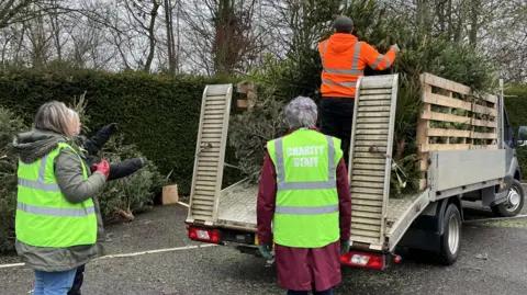 Tom Jackson/BBC A small van with an open back trailer is parked across the right hand side. Three people are stood on the concrete, wearing green Hi-Vis jackets, looking at the van. A man is stood on the van holding a green tree, and is wearing an orange Hi-Vis jumper. 