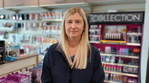 A woman is standing in a pharmacy. Her jacket says McGrath's Pharmacy. She has long blond hair.