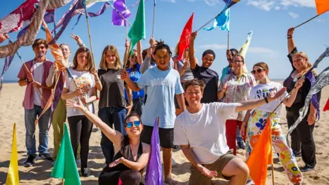 A group of young people on a sandy beach at Great Yarmouth. They are wearing summery clothes and are waving colourful flags and looking happy. The sky behind them is blue.