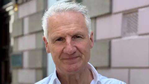 A man called Tom Binet. He is middle-aged and has trimmed white hair. He is wearing a white shirt and is stood in front of a building with pink bricks.