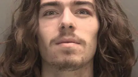 Merseyside Police A police mugshot of Connor Chapman, in his early 20s, who has long wavy brown hair, a brown moustache and a goatee beard and is staring into the camera with a slight smirk. 