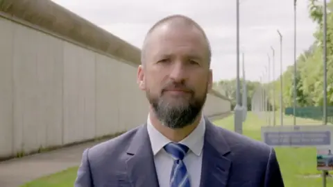 A man with very short hair and a beard is standing outside a prison wearing a blue suit. The prison wall is visible over his shoulders. 