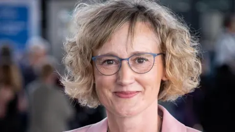 Welsh government Welsh language Commissioner Efa Gruffudd Jones. She has a blonde short curly bob with a side fringe. She wears grey glasses and her pink collar can be seen in the bottom of the frame. She has brown eyes and smiles at the camera.