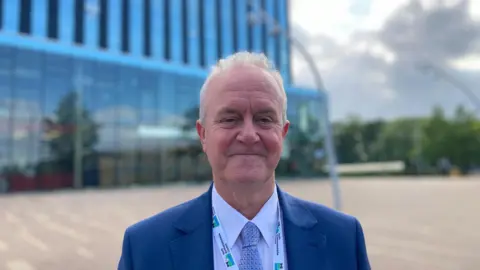 An older man with short white hair, smiling. He's wearing a navy blue suit jacket, with a white shirt and blue and white patterned tie. He has a North Northamptonshire Council lanyard. He's standing in front of a large modern building, with a grey cloudy sky behind him.