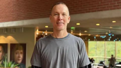  A man in a grey t-shirt smiles at the camera. He has short grey hair. There is a cafe behind her. There are large photos on the wall.