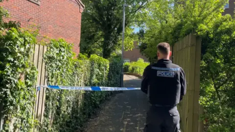 The back of a police officer in front of some blue police tape with a path behind the tape. The path goes between two fences with trees overhead.