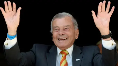PA Media Former Test umpire Harold 'Dickie' Bird at Yorkshire County Cricket Club. He holds his hands in the air. He is wearing a suit and a yellow and orange tie.