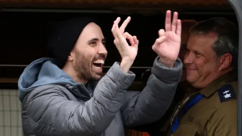 Reuters Tal Shoham, dressed in grey anorak, blue hoodie and black hat, smiles and gestures towards a crowd after being released by Hamas