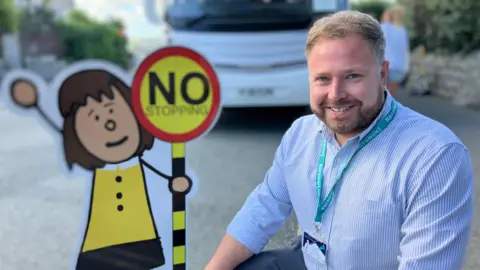 Trenance Learning Academy head teacher Matt Williams crouches down next to one of the yellow and black Parking Buddies signs. Matt is wearing a blue striped shirt and has a turquoise lanyard. A white bus is parked behind him.