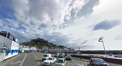 The seafront of a picturesque coastal town on Jersey. There is a street with houses and shops on with cars parked. There is a harbour and a castle in the background overlooking the town. 
