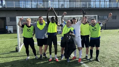 Bristol Hoteliers Association Seven footballers standing on a pitch in front of a net, wearing hi-vis jackets over their kit and waving their arms in the air in triumph. There is a sports centre building with a balcony stretching along its full length in the background.