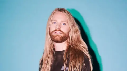 Sam Ryder A man with long blonde hair and a blonde/ginger beard staring into the camera