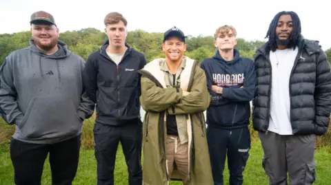 BBC Studios/Eddie Hutton-Mills Five men stand together by a lake, with trees behind it. They are all wearing coats and smiling at the camera.