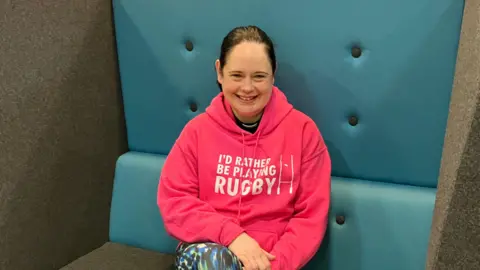 Reverend Beks Rothnie smiles at the camera. She is wearing a bright pink hoodie with "I'd rather be playing rugby" written on it.