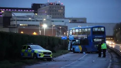 A blue double-decker bus is across a carriageway on an airport road and the door is hanging off. A police car is nearby on a verge with the back of the vehicle in a hedge and damage to the front of the marked vehicle. Two police officers with hi-vis jackets are standing near the bus. One is looking at a notebook and the other is talking on his phone.