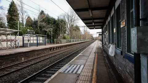 An image of a train station platform. It is surrounded by trees. It has two railway lines, two platforms and a brick waiting room with a small canopy to shelter the passengers. There is also a bike rack and a perspex shelter on the opposite platform.