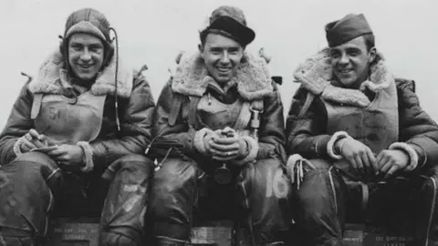 Three gunners, dressed in full World War Two uniform, sitting on the top of ammunition. They are all wearing carps or headgear, have their hands in their lap, are smiling to the camera, the image is black and white. 