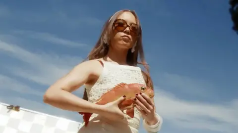 Laufey, framed against a sunny Californian sky, holds a red snapper while preparing to film a music video for her single Mad Woman